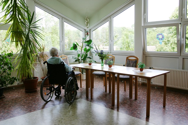 Eine alte Fraum sitzt im Rollstuhl allein an einem Tisch im Pflegeheim